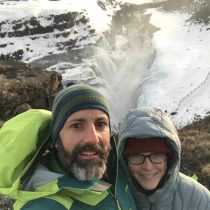 A man and woman in winter jackets and hats take a joyful selfie by a snowy waterfall after their Communicate with Impact & Influence coaching session outdoors.