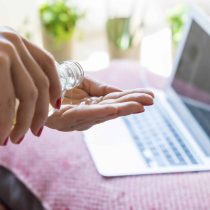 A person pours hand sanitizer onto their palm in front of an open laptop on a pink surface, likely preparing for Rejuvenating Face Yoga & Meditation, with green plants blurred in the background.