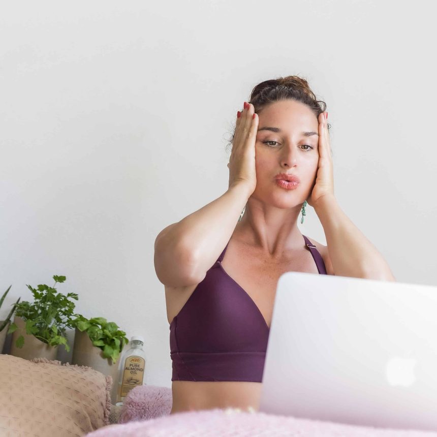A woman sits on a bed with her laptop, practicing techniques from Rejuvenating Face Yoga & Meditation. She holds her hands to her cheeks while plants and cushions decorate the background.