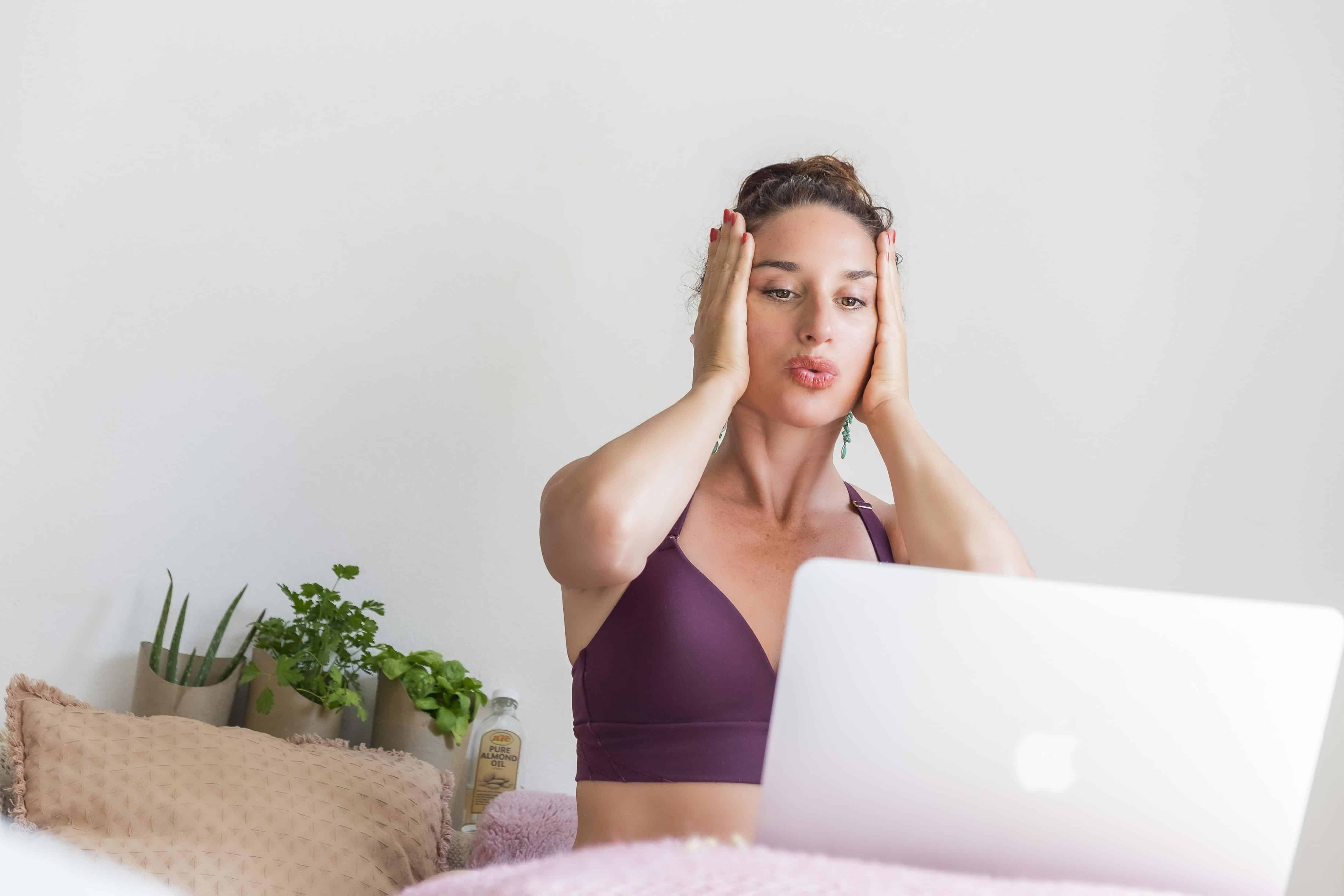 A woman sits on a bed with her laptop, practicing techniques from Rejuvenating Face Yoga & Meditation. She holds her hands to her cheeks while plants and cushions decorate the background.