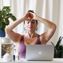 A woman sits at a desk with a laptop, eyes closed and hands in a yoga pose, possibly using techniques from the Rejuvenating Face Yoga & Meditation program. Plants and small bottles decorate the desk and background.