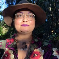 A person wearing glasses, a brown wide-brimmed hat, bright purple lipstick, and a colorful floral shirt stands outdoors with greenery and sunlight in the background. A Frida Kahlo tattoo is visible on their upper chest.