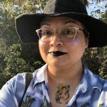 A person with glasses, black lipstick, glitter on their cheek, and a black wide-brim hat takes a selfie outdoors. They wear a blue palm tree shirt and have a colorful tattoo on their upper chest. Green foliage is in the background.
