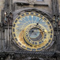 The Prague Astronomical Clock, featuring intricate gold and blue details, zodiac signs, Roman numerals, and surrounding statues, set into an old stone building facade.