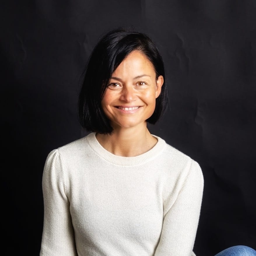 A woman with straight black hair and light skin, dressed in a white sweater, smiles against a plain black background, reflecting the transformative power of the 90-Day Journey to Deep Body Connection.