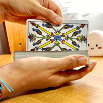A person prepares for an Online Intuitive Tarot Reading, shuffling a deck on a wooden table with the crossed wands card on top. Nearby, a white smiling object and wooden chair are visible in the background.