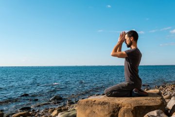 A man kneels on a large rock by a rocky shoreline, facing the ocean with hands pressed together in a meditation or prayer pose under a clear blue sky, seeking kundalini experiences and exploring what is tantra.