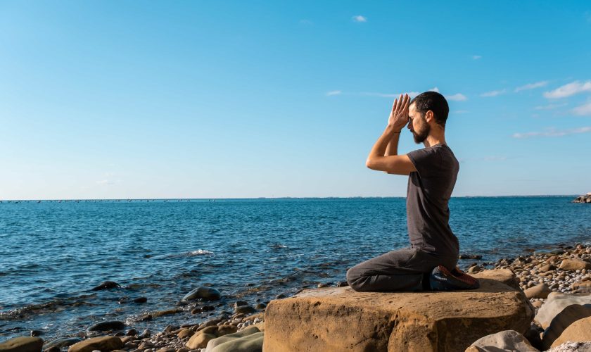 A man kneels on a large rock by a rocky shoreline, facing the ocean with hands pressed together in a meditation or prayer pose under a clear blue sky, seeking kundalini experiences and exploring what is tantra.