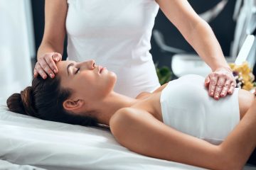 A woman lies on a massage table with her eyes closed as a therapist, acting as a reiki meditation coach, gently places one hand on her forehead and the other on her abdomen, creating a calm and relaxing atmosphere.