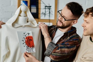 Two men smiling and holding a white hoodie with a red and black abstract design, in a bright room filled with shelves and art supplies—celebrating the journey of changing careers and discovering how to find your dream career.