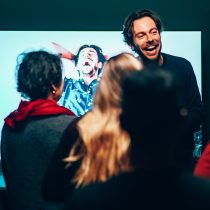 A man laughs in front of a TV showing a joyful person, while three colleagues watch in a dimly lit room—an ideal scene for incorporating Laughter As A Tool into your next quick team wellness activity.