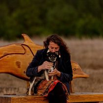 A woman explores grief with the Grief Exploration & Support Package, sitting on a wooden bench with a dolphin-shaped backrest and hugging a small black and white dog in her lap outdoors amid dry grass and trees.