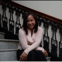 A woman with long dark hair, wearing a light pink top and black pants, sits smiling on a marble staircase with ornate black railings.