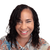 A woman with wavy, shoulder-length dark hair wearing a colorful patterned blouse smiles warmly against a plain white background.