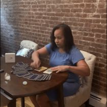 A woman in a blue dress sits at a round table, spreading tarot cards. The table also holds a small crystal, candle, and a card that says I CAN. She sits in front of a brick wall on a cushioned chair.