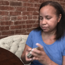 A woman in a blue shirt sits at a table shuffling a deck of cards. She has medium-length brown hair and is in front of a brick wall, with a patterned pillow behind her.