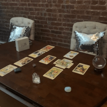 A wooden table with tarot cards, crystals, a crystal ball, and a box labeled I CAN, set between two chairs with shiny silver sequin pillows, against an exposed brick wall.