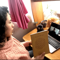 A woman in a pink sweater sits at a desk holding a book, looking at a laptop screen during a video call with two people. A window with pink curtains and potted plants are visible in the background.
