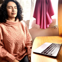 A woman with curly hair sits at a wooden desk with a laptop, eyes closed, wearing a pink sweater, appearing relaxed and meditative; sunlight streams through a window with pink curtains beside her.
