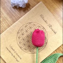 A crocheted pink tulip with green leaves rests on a book titled Akashic Records, featuring a geometric flower design on a wooden surface. A white crystal cluster is placed above the book.