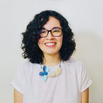 A person with curly dark hair and glasses smiles at the camera. They are wearing a white shirt and a statement necklace with blue and beige shapes, standing against a plain light-colored background.