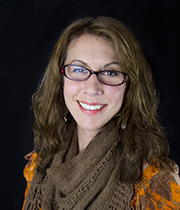 A woman with wavy brown hair, glasses, a brown scarf, and an orange top smiles warmly at the camera against a black background, embodying the warmth and comfort found in the Grief Exploration & Support Package.