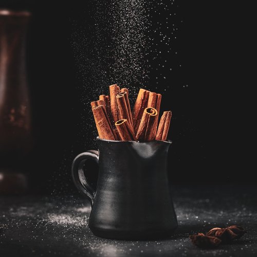 A black ceramic mug filled with upright cinnamon sticks sits against a dark background, with a gentle sprinkle of powder falling over the mug and a few spices scattered nearby.
