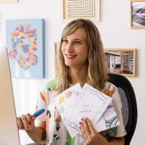 Flor de Valle, smiling at her computer during a virtual “Create a Greeting Card with Doodle Art” class, holding her illustrated cards with colorful artwork and photos on the wall behind her.