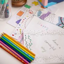 Get inspired for your next online team building art session with Create a Greeting Card with Doodle Art—colorful pens, markers, and a “Have a wonderful day” note decorate the white desk background.