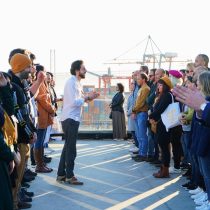 In Laughter As An Ally, a group stands in two lines outdoors for a team stress relief workshop. A man in the center gestures while speaking, as others listen attentively. Construction cranes can be seen in the background.