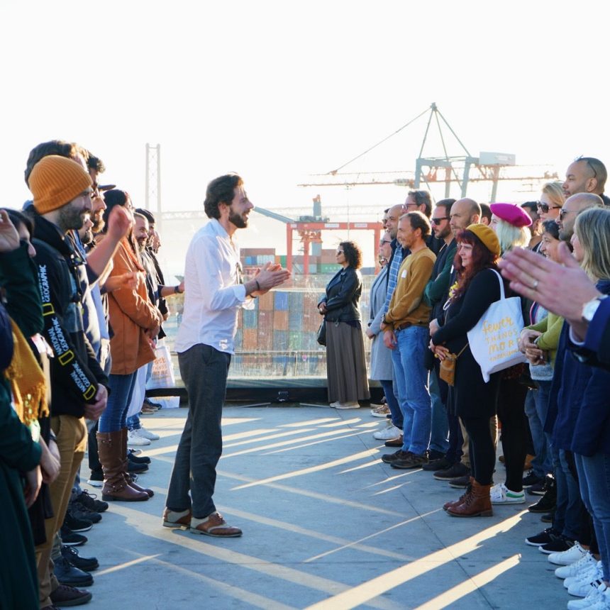 In Laughter As An Ally, a group stands in two lines outdoors for a team stress relief workshop. A man in the center gestures while speaking, as others listen attentively. Construction cranes can be seen in the background.