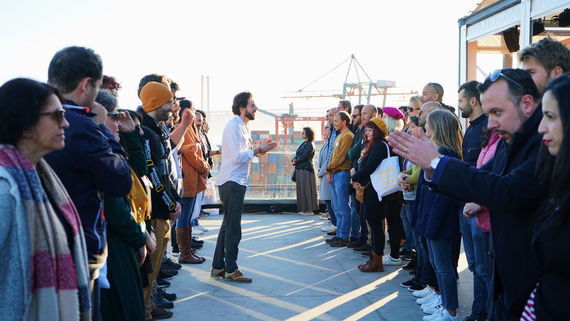 In Laughter As An Ally, a group stands in two lines outdoors for a team stress relief workshop. A man in the center gestures while speaking, as others listen attentively. Construction cranes can be seen in the background.
