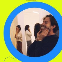 A group smiles and laughs at a team workshop. One man in the foreground touches his face and laughs. The image features a bright yellow background, blue circle, and Laughter As An Act of Consciousness at the top.