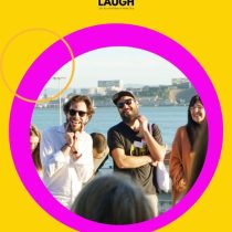 A group of smiling people stand outdoors by the water, framed by a bright pink circle and yellow background with FALL IN LAUGH—a perfect scene for team bonding with Laughter As An Act of Consciousness.