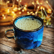 A blue ceramic mug of a frothy drink sits on rustic wood, surrounded by cozy fairy lights and foliage—the perfect scene inspired by our Virtual Warm Winter Drinks-Making Workshop.