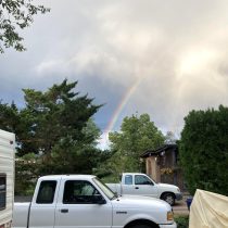 A faint rainbow arcs across a cloudy sky above trees and houses, with two white pickup trucks parked in a driveway, one partially covered by a tarp.