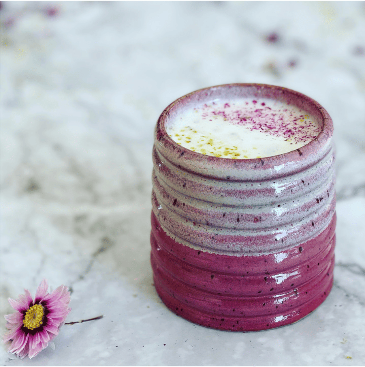 A pink and white ceramic cup with a creamy, frothed drink topped with pink and yellow powder sits on marble beside a daisy—perfect inspiration for your Virtual Drink-Making Team Building Workshop with Dancing Chef.