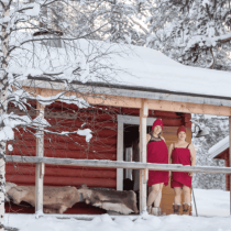 Two people in red towels and hats stand on a snowy porch, surrounded by winter scenery, embodying the spirit of Finnish Sauna Secrets from Lapland amid snow-covered trees and rooftops.