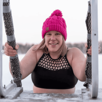 A smiling woman in a black swimsuit and bright pink knit hat emerges from icy water with snowy scenery, capturing the invigorating spirit of "Finnish Sauna Secrets from Lapland.