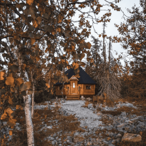 Inspired by Finnish Sauna Secrets from Lapland, a cozy wooden cabin with a dark roof nestles among orange-leaved trees on rocky ground, a gravel path winding nearby under a clear sky.