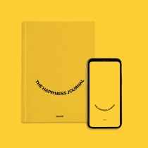 The Happiness Journal sits beside a smartphone showing its matching cover on a bright yellow background.