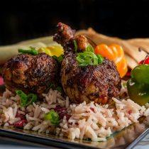 Cook with Jamaicans: Two seasoned, grilled chicken drumsticks served on rice with chopped green onions, garnished with whole green and orange peppers on a square plate. A cloth napkin sits in the background.