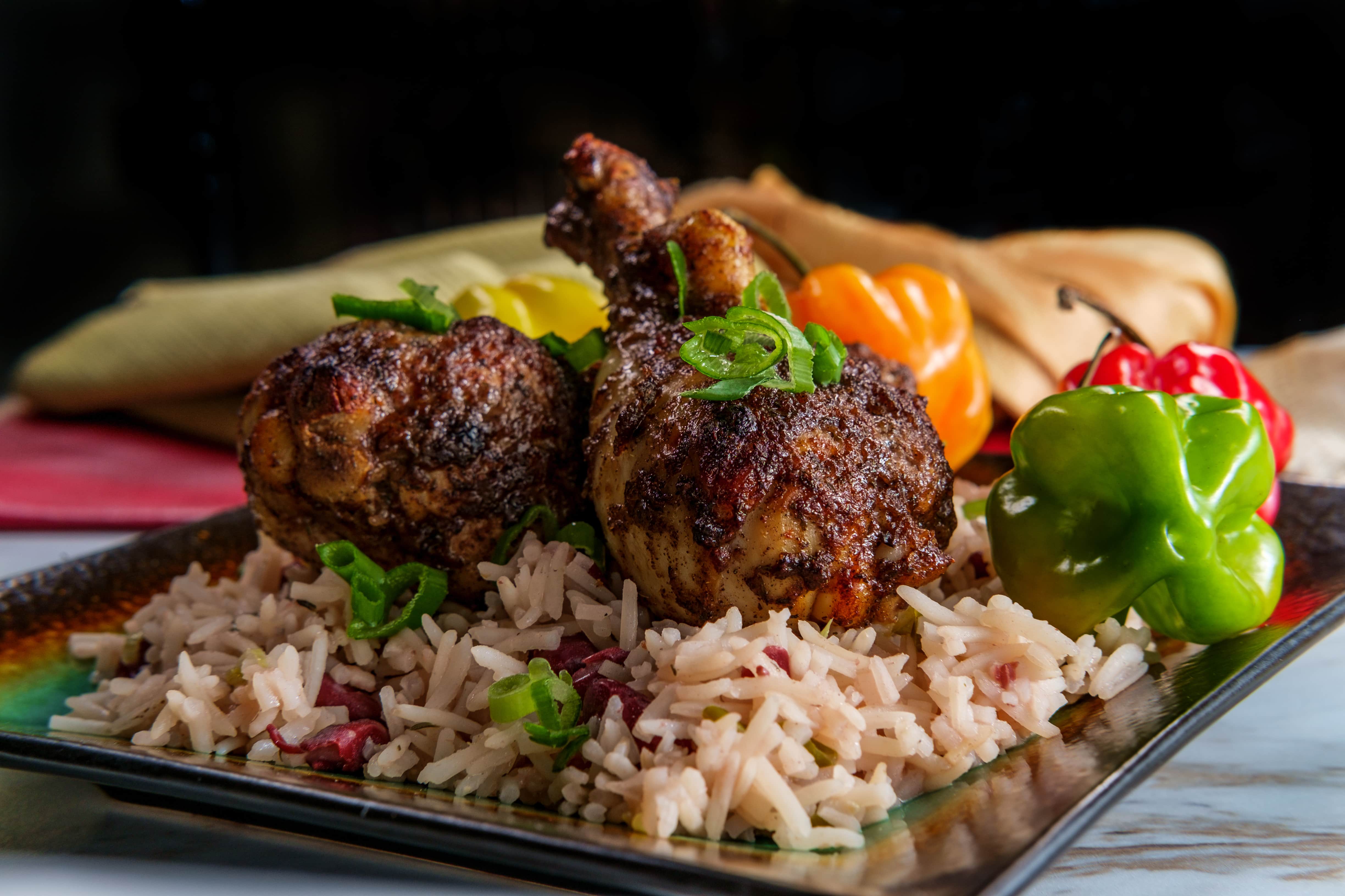 Cook with Jamaicans: Two seasoned, grilled chicken drumsticks served on rice with chopped green onions, garnished with whole green and orange peppers on a square plate. A cloth napkin sits in the background.