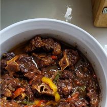 Cook with Jamaicans oxtail stew features tender meat, bones, and colorful bell peppers in a rich brown sauce, served on a kitchen counter.