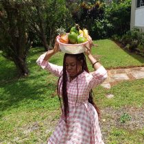 A woman in a pink and white checkered dress, featured by Cook with Jamaicans, stands on grass carrying a large metal bowl of mangoes and papayas on her head, with trees and a building in the background.
