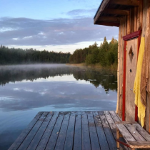 A wooden cabin with a yellow towel hanging outside sits beside a calm lake, with a dock leading to the water. Mist rises from the lake, and trees line the far shore under a cloudy sky.