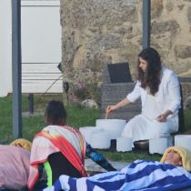 A woman leads a Sound Bath for Relaxation & Restful Sleep outdoors, playing crystal singing bowls as people rest on mats and blankets near a stone wall and grass, creating a peaceful atmosphere.