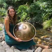 A woman in a green dress sits by a stream, smiling with a handpan drum in her lap. Surrounded by lush greenery, she enjoys the peaceful outdoors—an ideal scene for using Sound Bath for Relaxation & Restful Sleep.