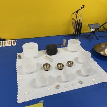 A white mat holds crystal and metal bowls, candles, and stones—creating a serene space for your Sound Bath for Relaxation & Restful Sleep. Musical instruments rest on a blue floor with a bright yellow wall backdrop.