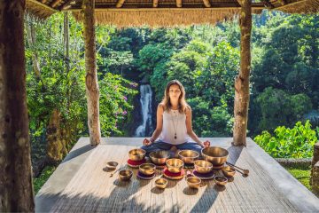A woman sits cross-legged on a gazebo platform surrounded by singing bowls, meditating and enjoying a sound bath with a lush forest and waterfall in the background.